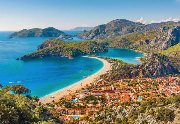 A coastal town with red-roofed buildings stretches along a sandy beach, while a vibrant blue bay and lush, green hills provide a scenic backdrop under a clear sky.